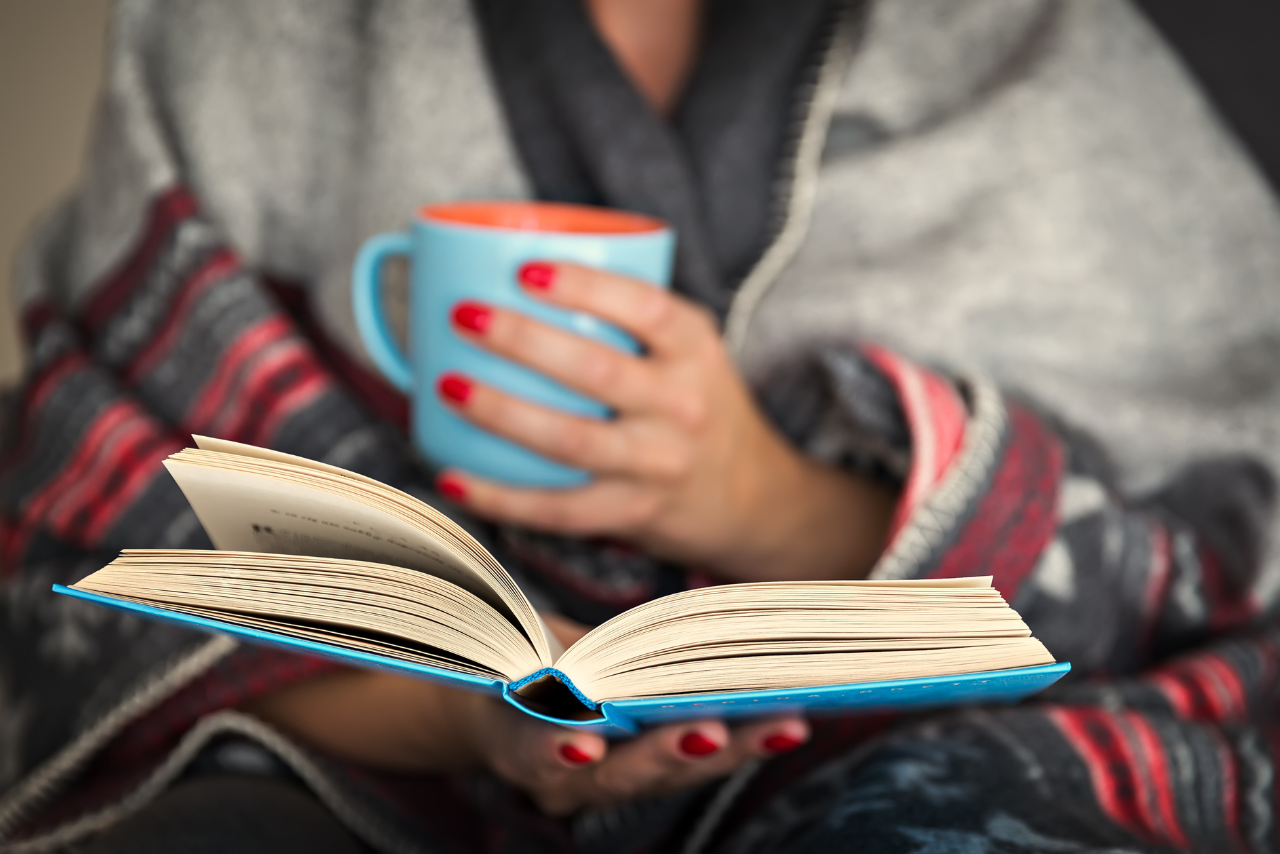 Cozy reading scene with a person wrapped in a warm blanket, holding a blue mug and an open book, evoking relaxation and comfort. Ideal for a winter reading ambiance or book lover lifestyle concept.