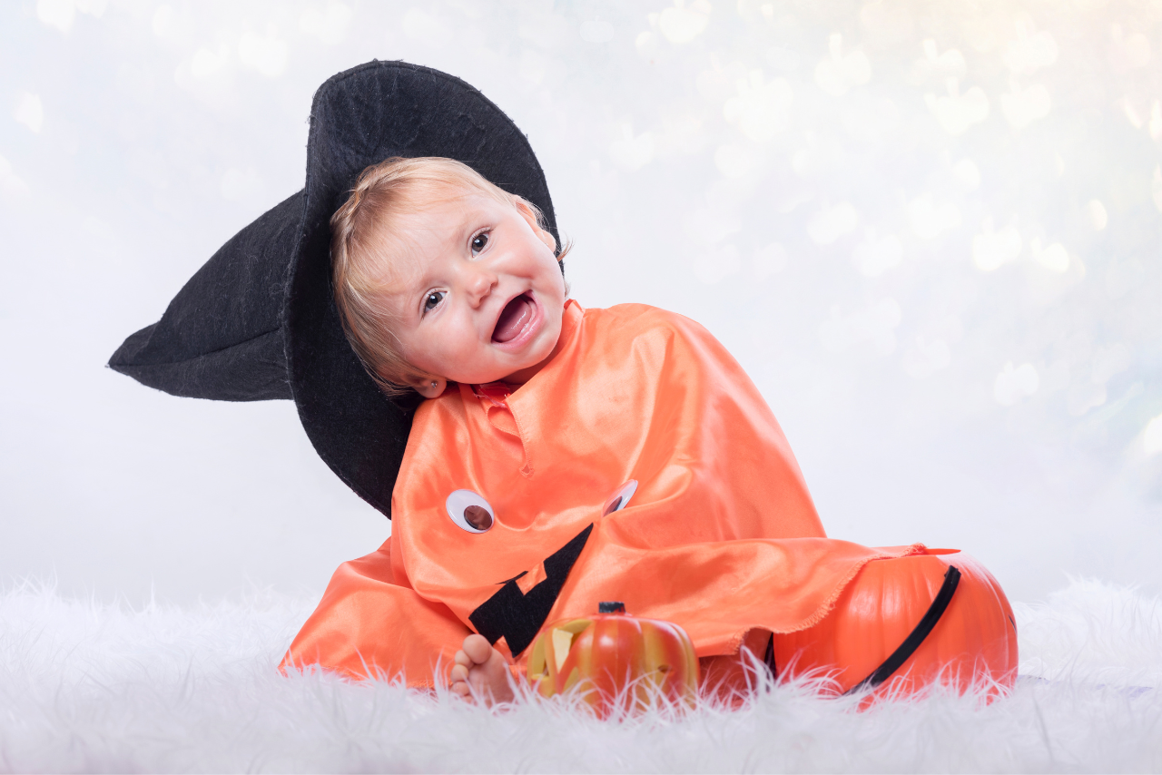 Cute baby dressed in an orange pumpkin costume with a black witch hat, smiling and sitting on a fluffy white surface, perfect for Halloween.