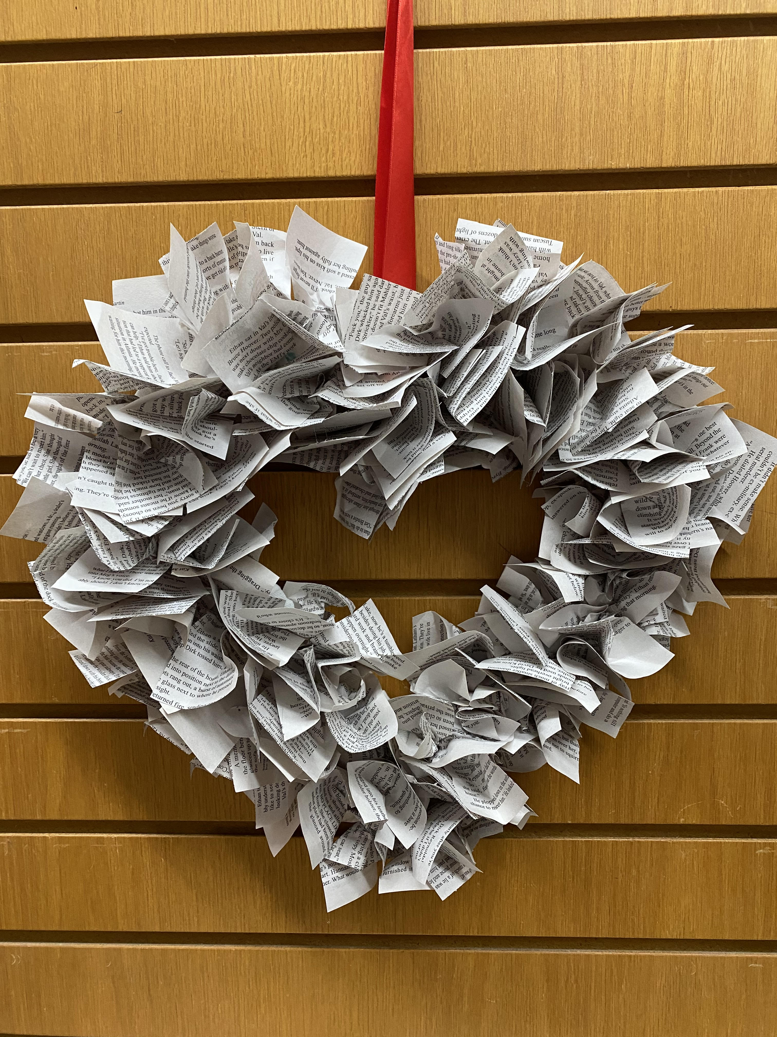 Brown slat wall with a heart shaped wreath with book pages curled and glued to the heart shape.  A red ribbon is tied at the top