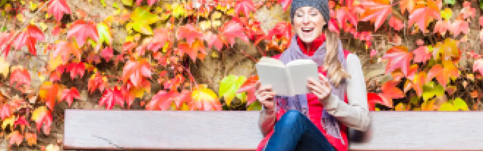 Woman with grey hat and red vest reading a book on a bench with colorful fall foliage behind her